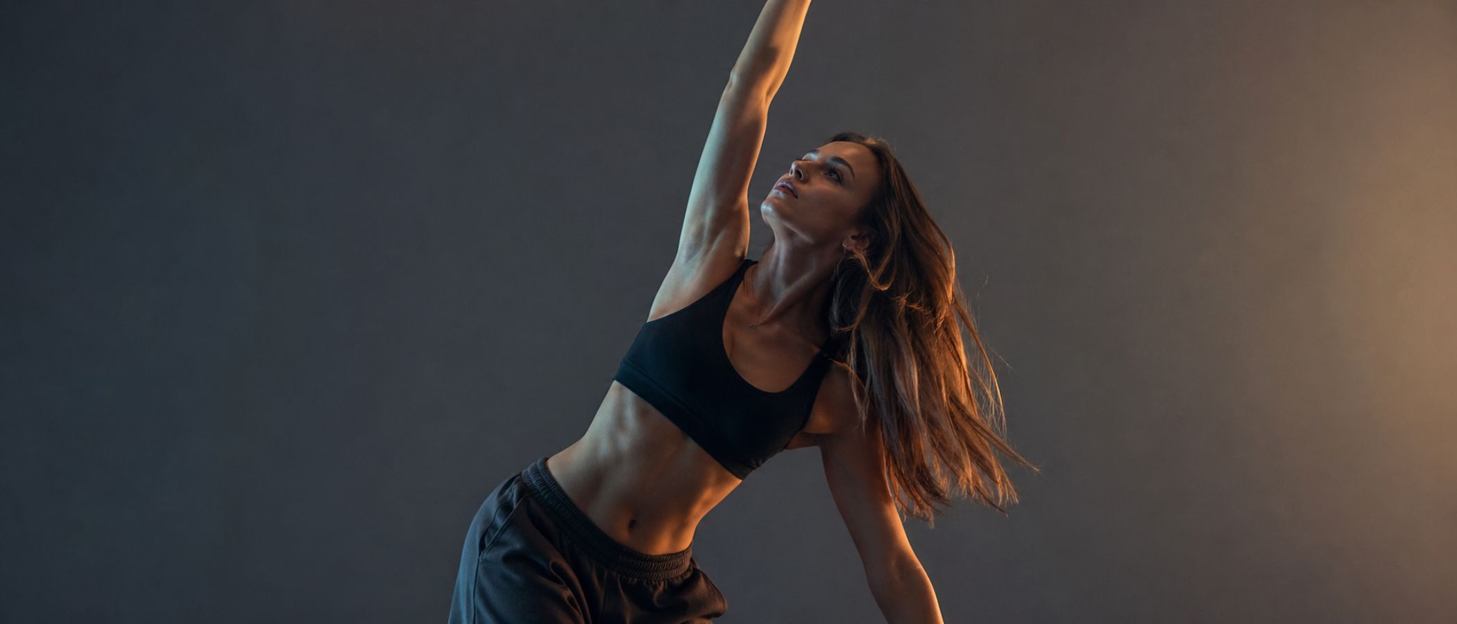 Woman in a black sports bra and shorts stretches with her right arm raised overhead against a dark gradient backdrop.