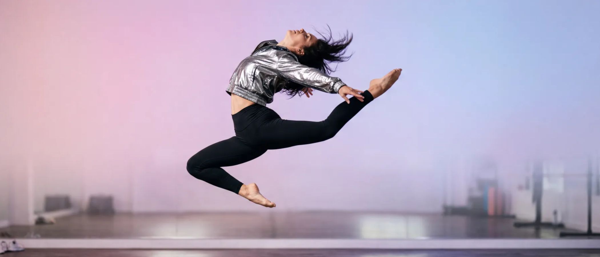 Dancer in a silver jacket and black leggings launches into a high leap midair against a pastel studio backdrop.