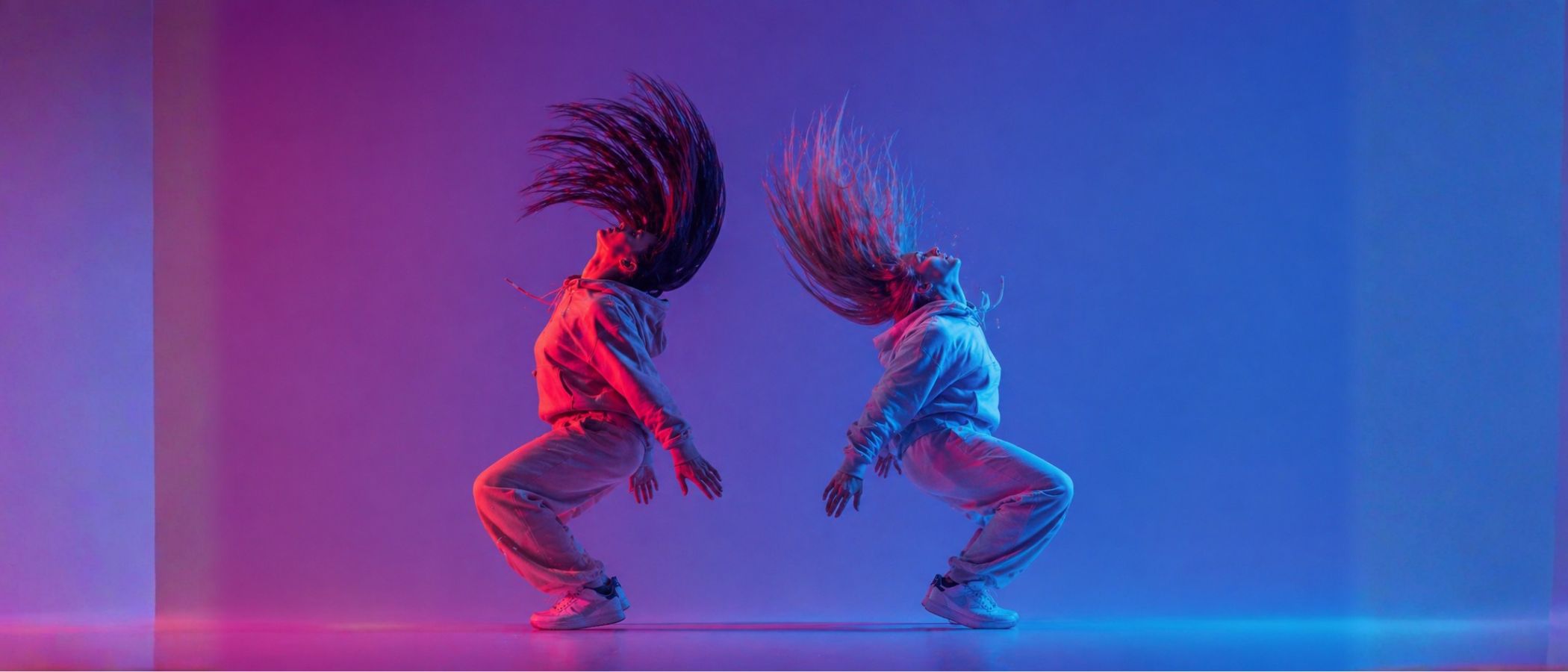 Two dancers in light outfits jump back-to-back with hair streaming, under neon pink and blue lighting.