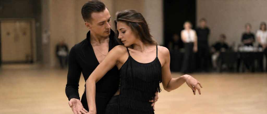 Couple dancing in black outfits: man behind woman, performing a ballroom step in a studio with onlookers in the background.