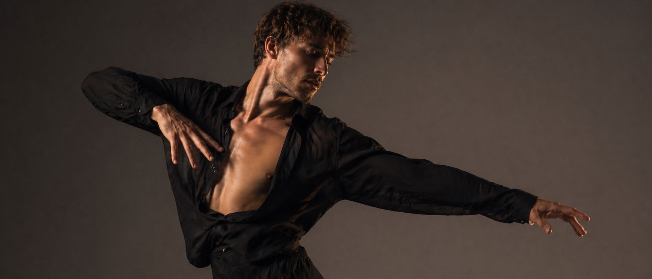 Male dancer in a black shirt performs a dramatic, extended-arm pose against a dark studio backdrop.