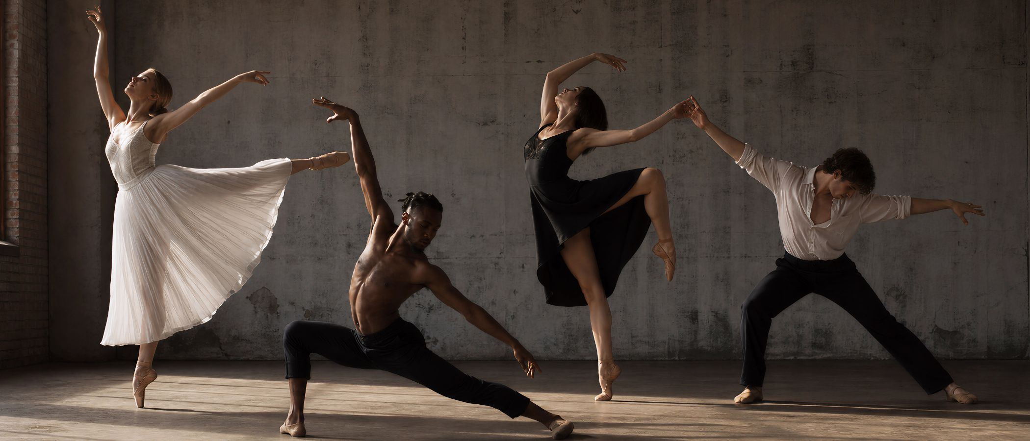 Five dancers perform a contemporary routine in a dim, industrial-studio space, each in expressive, extended poses across the room.