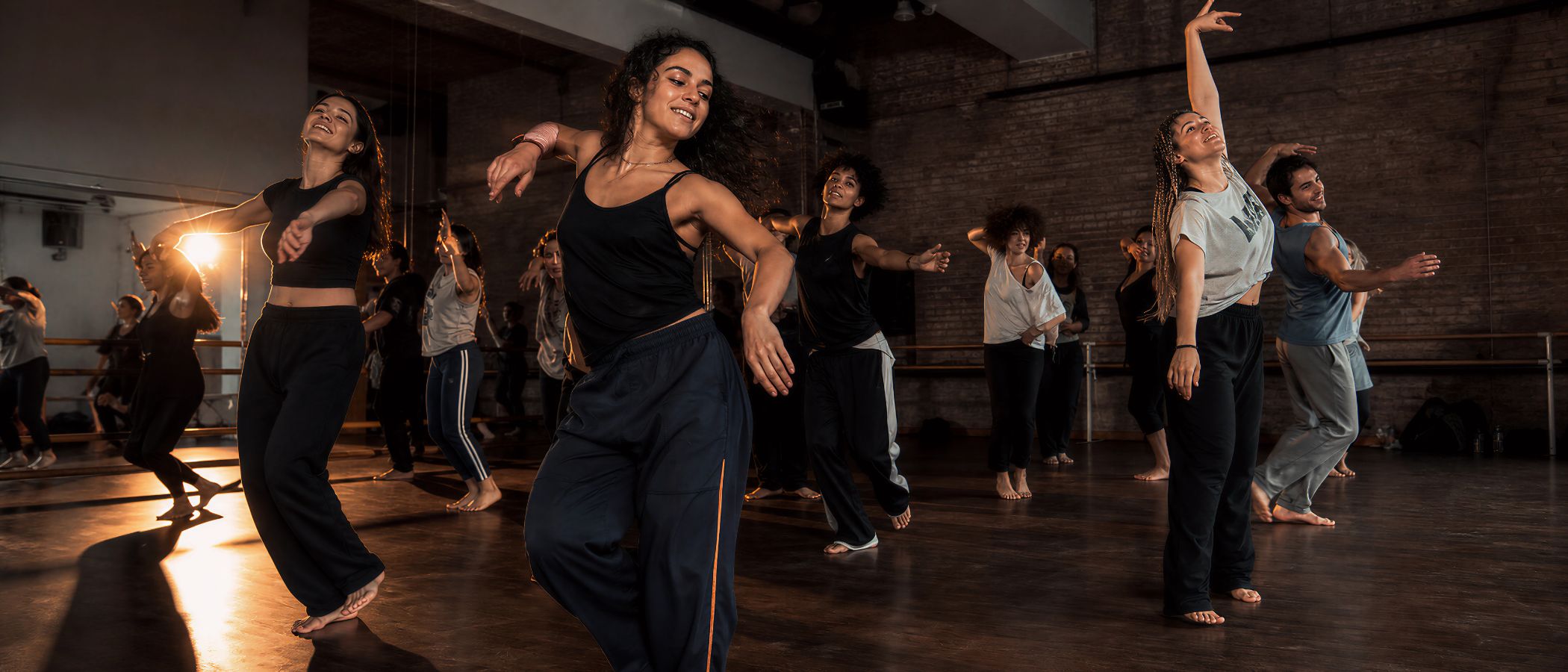 Diverse group of dancers in a studio, barefoot and mid-movement during a class with brick walls and mirrors.