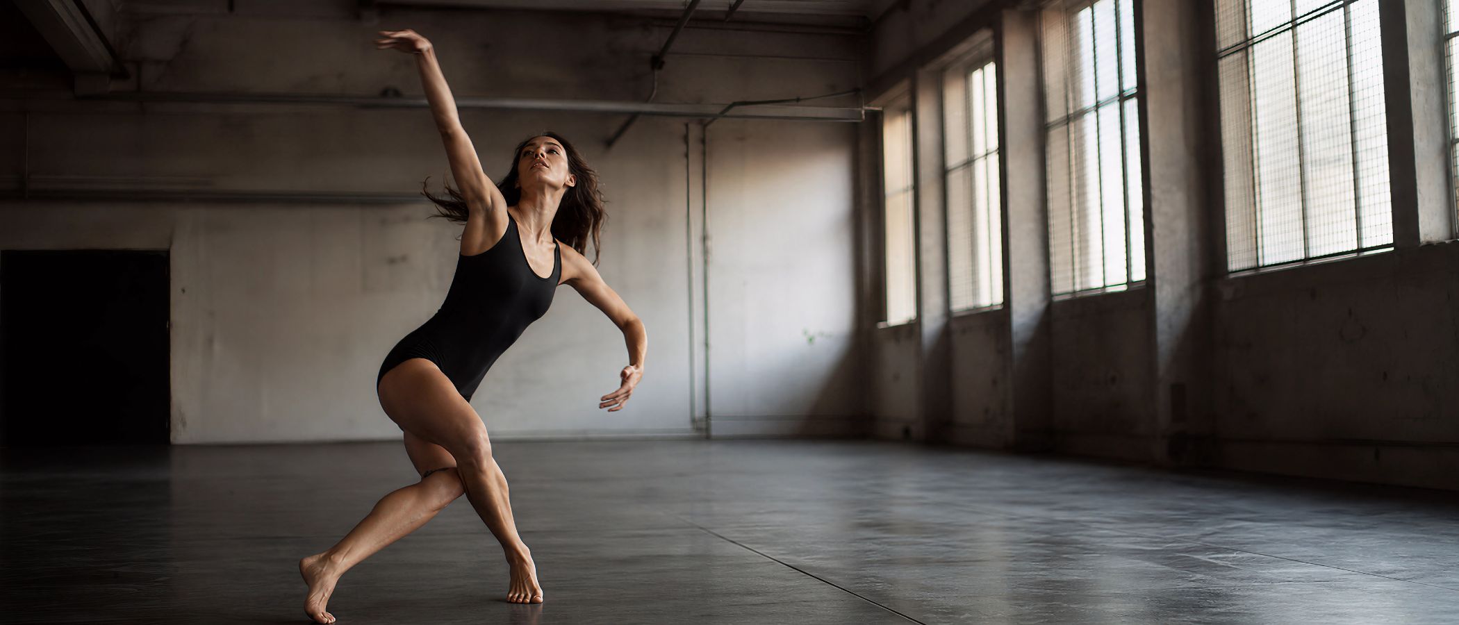 Female dancer in a black leotard performing a dynamic pose in an empty industrial studio with large windows.