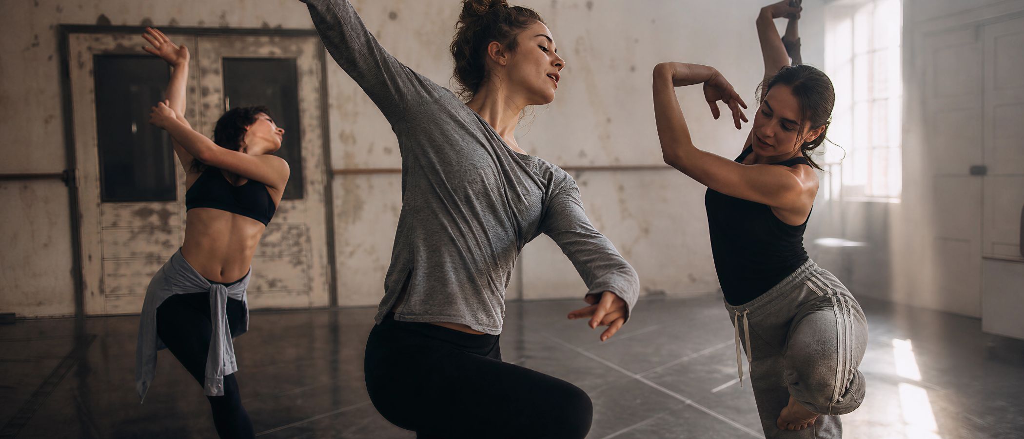 Three women dance in a sunny, industrial-style studio, performing expressive moves with arms raised and one leg lifted.