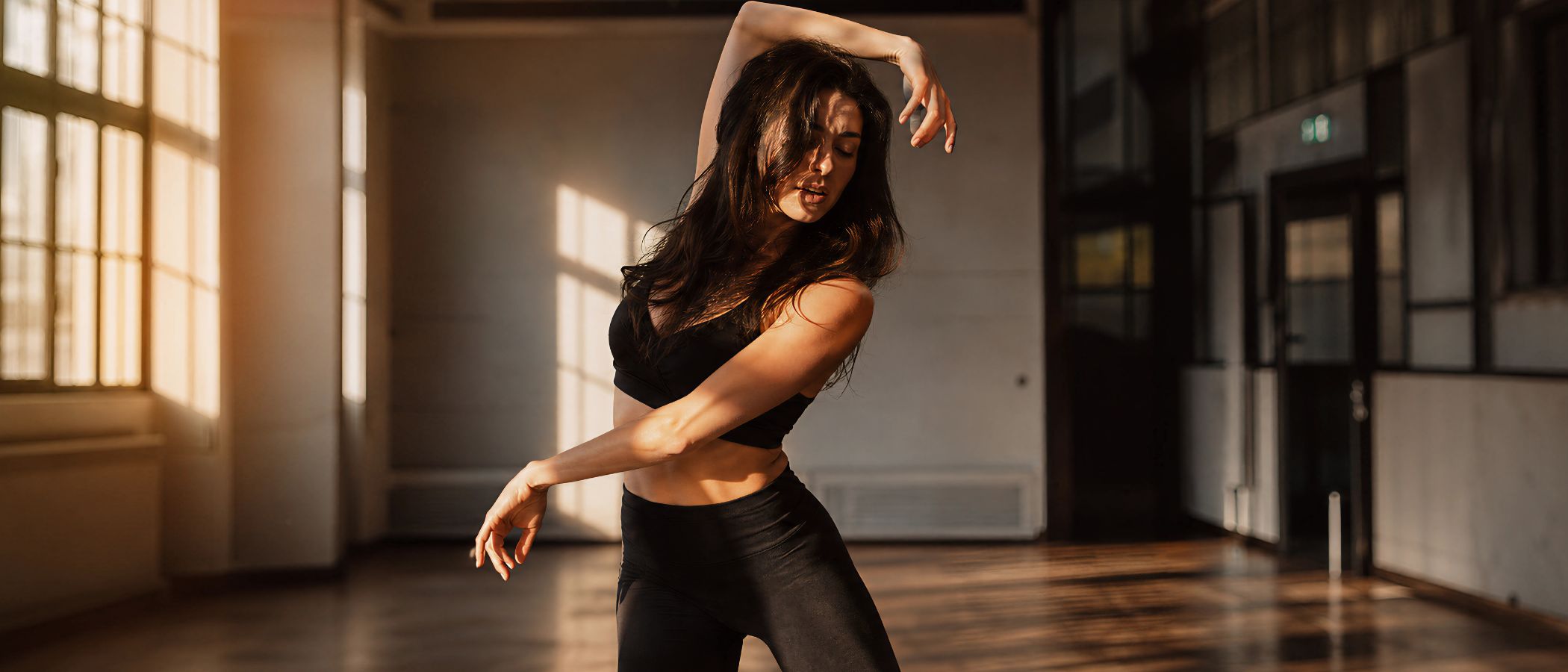 Woman wearing a black sports bra and leggings dancing in a sunlit studio, arms lifted and eyes closed.