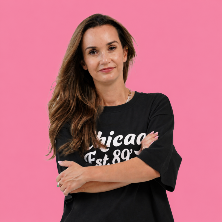 Portrait of a woman with long wavy hair, wearing a black t-shirt that says 'Chicago Est. 89' against a pink background, arms crossed and a slight smile.