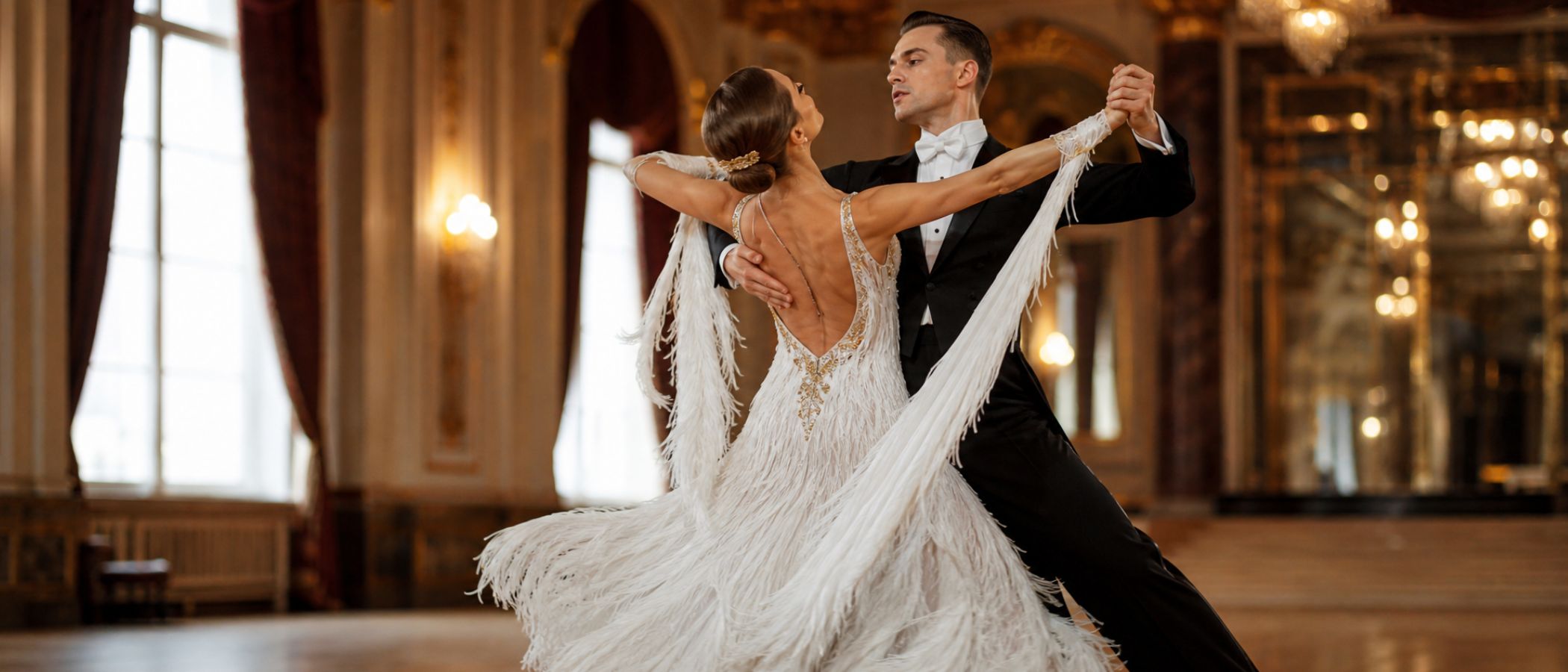 Couple ballroom dancing in a luxurious, gold-lit hall; woman in a white fringe gown and man in a black tuxedo hold a graceful pose with intertwined arms.